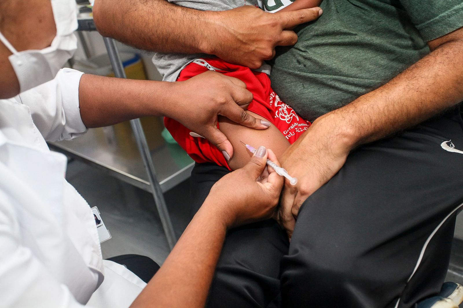 Profissional da saúde aplicando vacina na perna de uma criança que está sentada no colo do pai.